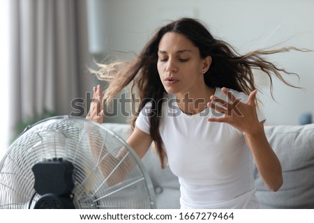 Millennial Caucasian girl sit on sofa in living room breathe fresh air from floor ventilator suffer from summer heat at home, young woman feel hot use fan at home struggle with lack of air condition Royalty-Free Stock Photo #1667279464