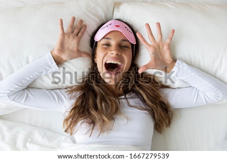 Top view portrait of overjoyed Caucasian millennial girl in sleeping mask look at camera lying in white cozy bed feel happy in morning, smiling young woman have fun posing in comfortable bedroom