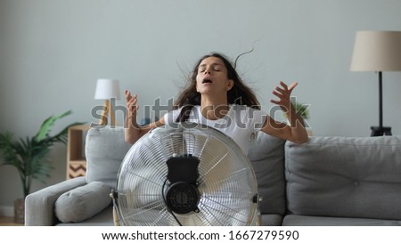 Young Caucasian woman sit on couch in living room breathe fresh air from floor fan suffering from lack of conditioner, millennial girl use ventilator struggle with home heat, summer hot weather Royalty-Free Stock Photo #1667279590
