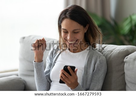 Excited millennial girl sit on couch in living room look at cellphone screen feeling overjoyed euphoric winning lottery online, happy young woman triumph get pleasant smartphone message or text Royalty-Free Stock Photo #1667279593