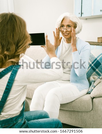 Little Granddaughter Making Videos Or Photos Of Her Beautiful Grey Haired Modern Grandmother Adjusting Her Eye Glasses And Looking At Camera, Video Concept, Toned Image