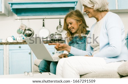 Mature Grandmother And Her Little Granddaughter Sitting Together On Sofa And Looking On Screen Of Laptop Computer Smiling Girl Showing Her Granny Some Pictures Or Photos, Toned Image
