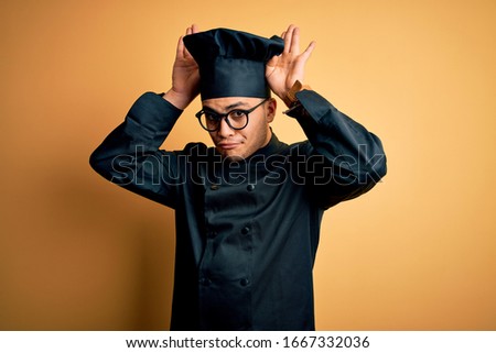 Young brazilian chef man wearing cooker uniform and hat over isolated yellow background Doing bunny ears gesture with hands palms looking cynical and skeptical. Easter rabbit concept.