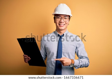 Young handsome chinese engineer man wearing safety helmet holding clipboard very happy pointing with hand and finger