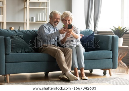 Happy middle-aged 50s husband and wife sit rest on couch in living room have fun making self-portrait picture on cell, excited elderly 60s couple laugh taking selfies using modern smartphone together