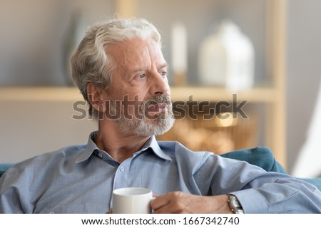Thoughtful elderly 60s husband relax on couch at home drink tea pondering or mourning, pensive middle-aged old man sit rest on sofa in living room look in window distance thinking or dreaming Royalty-Free Stock Photo #1667342740