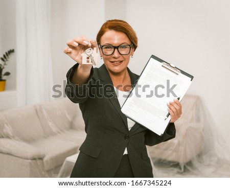 realtor propose to sign an agreement for apartment rent, camera focused on hand keeping keys. Woman in glasses and busines suit standing in the room with keys in one hand and agreement in another.