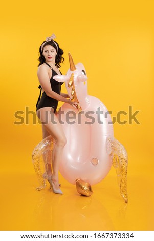 Young adult brunette girl with pin-up hairstyle in a black swimsuit posing with a beach inflatable unicorn toy on a yellow background