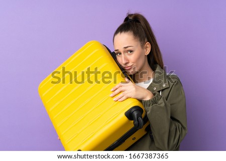Young brunette woman over isolated purple background in vacation with travel suitcase and unhappy