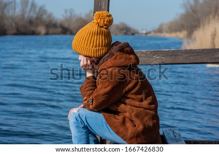 A girl in a light sheepskin coat stands on the bank of the river on a sunny day. Girl 9 years old in a sheepskin coat by the river. Girl 9 years old or 10 years old. Nature, child, schoolgirl.Teenager