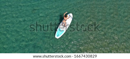 Aerial drone ultra wide photo of fit woman enjoying SUP or Stand Up Paddle board in shallow exotic bay with emerald sea