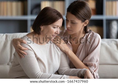 Young compassionate woman embracing shoulder of unhappy stressed middle aged mommy, comforting her at home. Kind grownup daughter supporting caring of older mature mother, sitting together on couch. Royalty-Free Stock Photo #1667437960