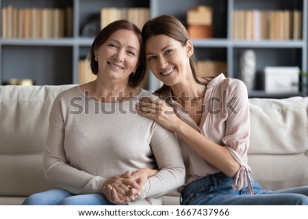 Portrait of happy grownup daughter touching shoulder of pleasant smiling old mature mother, relaxing together on couch. Affectionate two generations family looking at camera, posing for photo at home.