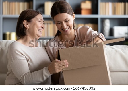 Happy older mature mommy sitting on couch with smiling grownup child, unpacking parcel together at home. Excited different female generations clients unboxing ordered goods from internet store. Royalty-Free Stock Photo #1667438032