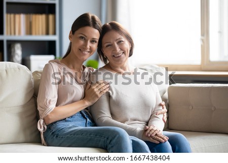 Portrait of smiling young woman cuddling affectionate older mother, sitting together on cozy sofa at home. Attractive happy different generations family relaxing in living room, looking at camera.