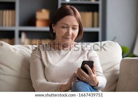 Pleasant senior older lady resting on couch, using applications on smartphone. Happy old mature woman chatting in messenger or social network with friends or children, shopping in internet store.