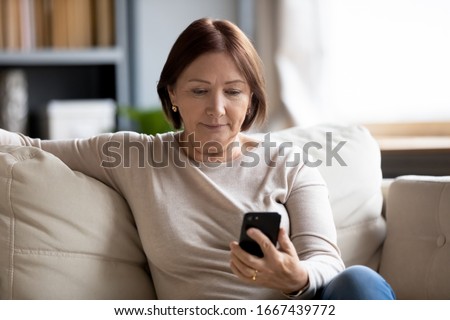 Worried middle aged woman sitting on couch, looking at mobile phone screen. Serious senior mature mother waiting for response from grownup children, chatting in social network online via smartphone. Royalty-Free Stock Photo #1667439772