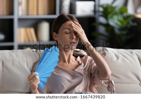Head shot stressed unhealthy young woman using paper fan, suffering from heat stroke at home. Unhappy 30s lady sit on sofa, feeling high temperature indoors without air conditioning in living room. Royalty-Free Stock Photo #1667439820