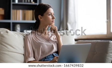 Distracted from work worried young woman sitting on couch with laptop, thinking of problems. Pensive unmotivated lady looking at window, feeling lack of energy, doing remote freelance tasks at home. Royalty-Free Stock Photo #1667439832