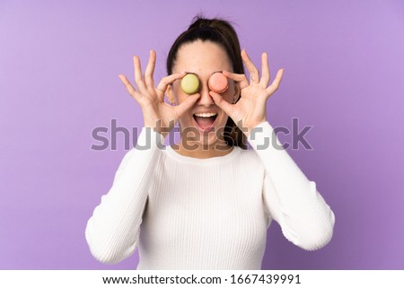 Young brunette woman over isolated purple background holding colorful French macarons with surprised expression