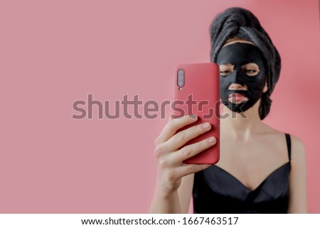 Young woman apply black cosmetic fabric facial mask and phone in hands on pink background. Face peeling mask with charcoal, spa beauty treatment, skincare, cosmetology. Close up.
