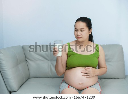 Young Asian pregnant woman holding  glass of milk. Healthy living. Her sitting in liveing room. Nutrition and diet during pregnancy.