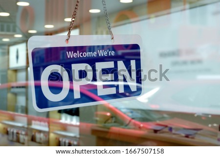 A 'Welcome We're Open' sign hanging on a glass entrance door of a shop selling electronic gadgets. Store with open for business signage welcoming customers to shop during its operating hours.