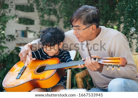 Old man with gray hair teach his grandson play the guitar in the backyard of his house.