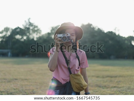 Happy beautiful young adventurous woman in casual clothes with hat, camera and map is taking photo with old vintage camera in forest while walking and hiking for camping on epic mountain.