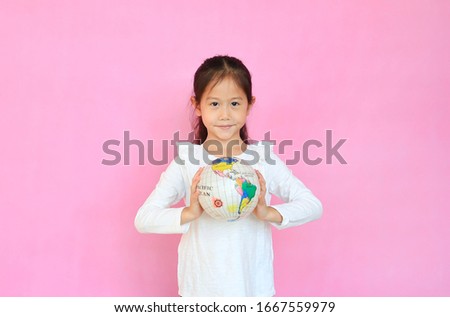 Cute asian little kid girl holding a globe with looking at camera isolated on pink background. Portrait of child with the earth globe sphere. Focus at children face.