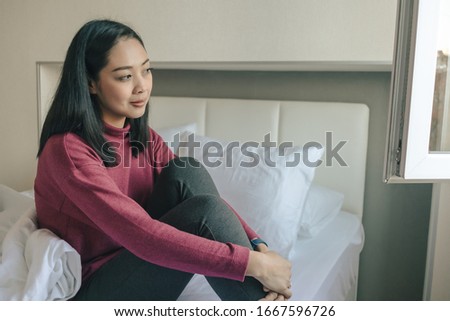 Asian woman is sitting on the bed and looking out to the window in the morning.
