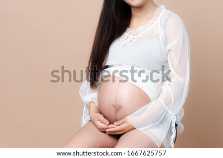 Pregnant woman carry her belly with hands siting on beige background