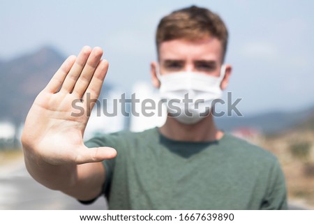 Guy, young man in protective sterile medical mask on his face looking at camera outdoors, on asian street show palm, hand, stop no sign. Air pollution, virus, Chinese pandemic coronavirus concept.
