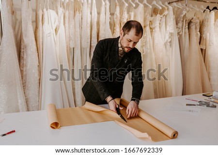 Young serious tailor with beard looking on drawing near wooden table with threads in amazing atelier with antique furniture and mannequin