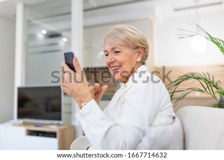 Mature woman with mobile phone on her hands sitting in room and sending messages to her friends and family. Senior woman texting on her mobile phone