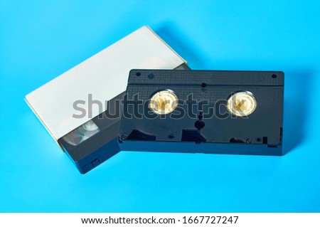Black old vhs video cassette in white cardboard packaging on blue desk. Concept of 90s. Close-up