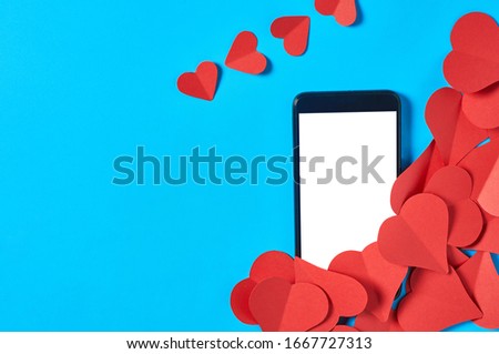 Black smartphone with isolated white screen for text, picture, photo and other graphics near heap of red paper hearts on blue table. Secrets or forbidden love concept. Space for text. Top view