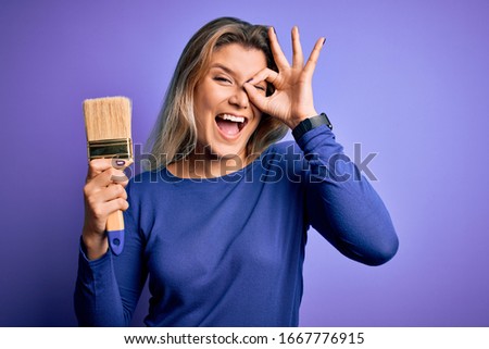 Young beautiful blonde painter woman painting using paint bush over purple background with happy face smiling doing ok sign with hand on eye looking through fingers