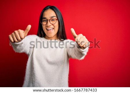 Young beautiful asian woman wearing casual sweater and glasses over red background approving doing positive gesture with hand, thumbs up smiling and happy for success. Winner gesture.
