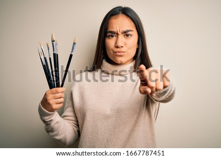Young beautiful artist asian woman holding paint brushes standing over white background pointing with finger to the camera and to you, hand sign, positive and confident gesture from the front