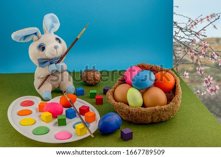 Colored easter eggs in the nest of straw. Gray plush rabbit with a palette with a brush paints Easter eggs. Multi-colored cubes around. Green grass carpet and blue background.