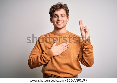 Young blond handsome man with curly hair wearing casual sweater over white background smiling swearing with hand on chest and fingers up, making a loyalty promise oath