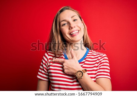 Young beautiful blonde woman wearing casual striped t-shirt over isolated red background cheerful with a smile of face pointing with hand and finger up to the side with happy and natural expression