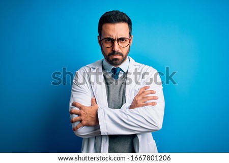 Young handsome doctor man with beard wearing coat and glasses over blue background skeptic and nervous, disapproving expression on face with crossed arms. Negative person.