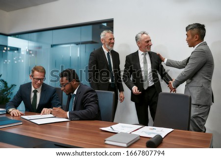 Two happy mature businessmen in classic wear shaking hands while having a multi-ethnic meeting in the modern office. Business concept. Business meeting. Partnership