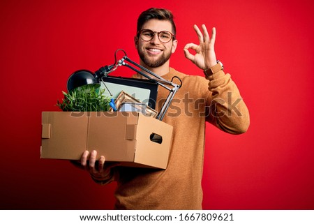 Young blond worker man with beard and blue eyes fired holding cardboard box doing ok sign with fingers, excellent symbol