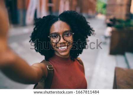 Young cheerful African American female in glasses dressed in casual clothes looking at camera with happy smile while standing in street at daytime