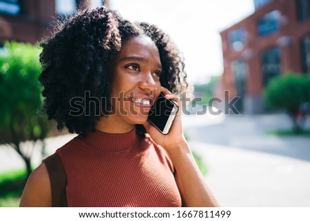 Smiling cheerful white toothed ethnic lady in casual apparel making call on mobile phone in street in summer in city with green trees