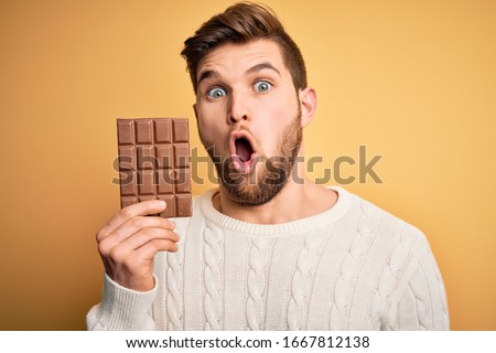 Young blond man with beard and blue eyes holding chocolate bar over yellow background scared in shock with a surprise face, afraid and excited with fear expression