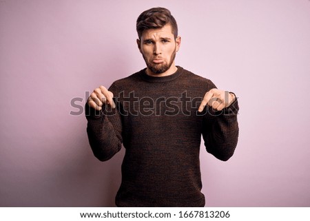 Young blond man with beard and blue eyes wearing casual sweater over pink background Pointing down looking sad and upset, indicating direction with fingers, unhappy and depressed.